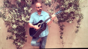 A man holding an acoustic guitar in front of some bushes.